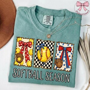K&ouml;nnte beinhalten: T&uuml;rkisfarbenes Comfort Colors T-Shirt mit Softball-Motiv. Das Design umfasst drei gerahmte Bilder von Softb&auml;llen, Handschuhen und Schleifen. Der Text "SOFTBALL SEASON" ist in fetter, wei&szlig;er Schrift gedruckt.