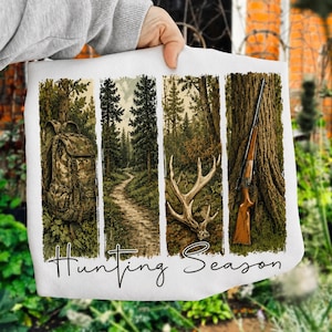 May include: A white sweatshirt featuring four vertical panels with illustrations of a camouflage backpack, a forest path, deer antlers, and a rifle leaning against a tree. The text Hunting Season is printed in black cursive script at the bottom.