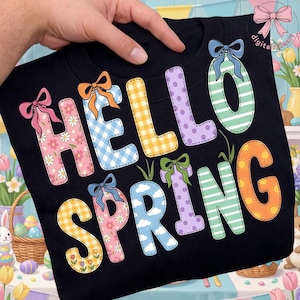 Könnte beinhalten: Schwarzes Sweatshirt mit dem Schriftzug "HELLO SPRING" in bunten Buchstaben im Patchwork-Stil. Die Buchstaben sind mit Blumen-, Vichy- und Wolkenmustern verziert, und jeder Buchstabe hat eine Schleife. Das Sweatshirt wird von einer Hand gehalten.