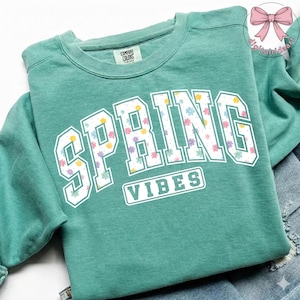 May include: A teal green sweatshirt featuring the text "SPRING VIBES" in large white block letters. The letters are filled with a floral pattern of pink, yellow, and purple flowers. The sweatshirt has a crew neck and long sleeves.