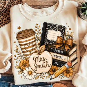 May include: A cream-colored crewneck sweatshirt featuring a graphic print of a striped coffee cup, a notebook, a stack of three books labeled TEACH, INSPIRE, and LOVE, a pencil, and an apple with the text Mrs. Smith, surrounded by floral accents.