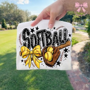 May include: White t-shirt with a softball theme. The design includes the word "SOFTBALL" in black, a yellow bow, a baseball bat, a brown glove, yellow softballs, and black stars. The design is perfect for softball players.