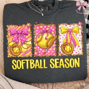 Könnte beinhalten: Dunkelgraues Sweatshirt mit Softball-Motiv. Das Design zeigt rosa und gelbe Schleifen, Softbälle, einen Baseballhandschuh und einen Schläger. Der Text "SOFTBALL SEASON" ist in gelben Blockbuchstaben.