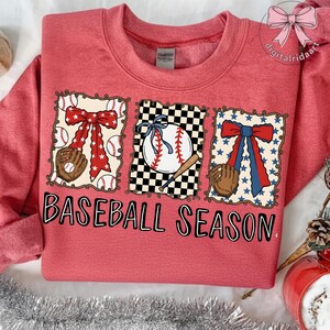 May include: Red sweatshirt with a baseball-themed design. The design includes a baseball, glove, and bow, with the words "BASEBALL SEASON" in black block letters.