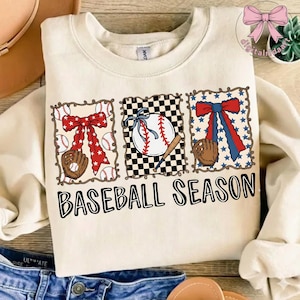 May include: Cream-colored sweatshirt with a baseball-themed design. The design includes a baseball, bat, and glove, along with the text "BASEBALL SEASON". The graphics are framed with a decorative border and bows.