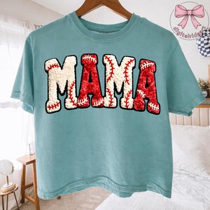 May include: A light blue t-shirt with the word "MAMA" in large, textured letters. The letters are designed to resemble baseballs, with red stitching on a white background. The shirt has short sleeves and a crew neck.