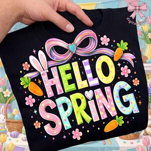 Może przedstawiać: Czarna bluza z kolorową grafiką "Hello Spring". Projekt zawiera uszy królika, marchewki, kwiaty i tęczową wstążkę. Tekst jest w pastelowych kolorach, idealny na Wielkanoc lub wiosnę.