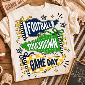 Puede incluir: Una camiseta color crema con tres gráficos en forma de banderín. El banderín azul superior dice FOOTBALL, el verde central dice TOUCHDOWN con líneas de campo, y el banderín amarillo a cuadros inferior dice GAME DAY, decorado con estrellas.