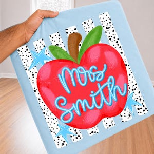 May include: A light blue fabric square featuring a large red apple graphic with green leaves and a brown stem. The apple is centered over vertical white stripes with black speckles and light blue stars. The text Mrs. Smith is written in blue.