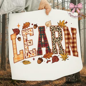 Puede incluir: Un letrero blanco con la palabra "LEARN" en letras grandes y decorativas. Las letras presentan varios patrones, incluyendo cuadros escoceses, estampado de leopardo y diseños de hojas de otoño. Hay bellotas y hojas dispersas alrededor de las letras.