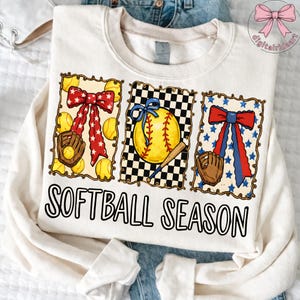 K&ouml;nnte beinhalten: Cremefarbenes Sweatshirt mit einem "Softball Season" Aufdruck. Die Grafik zeigt drei gerahmte Designs mit Softball-Elementen: Schleifen, Handschuhe, Softb&auml;lle und einen Baseballschl&auml;ger. Die Schleifen sind rot, wei&szlig; und blau.