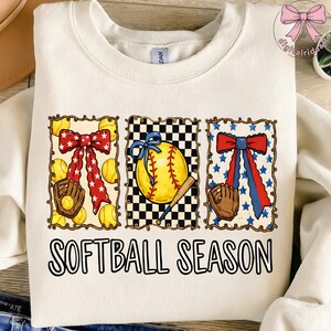 K&ouml;nnte beinhalten: Cremefarbenes Sweatshirt mit einem Softball-Motiv. Das Design umfasst drei gerahmte Bilder von Softb&auml;llen, Schleifen und Handschuhen. Der Text "SOFTBALL SEASON" ist in schwarzen Blockbuchstaben auf der Unterseite des Shirts aufgedruckt.