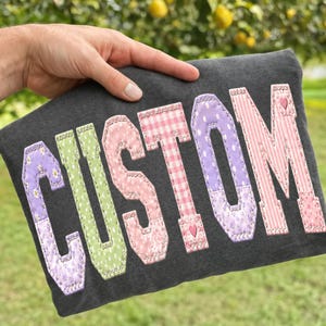 Può includere: Un articolo in tessuto grigio scuro con la parola CUSTOM in grandi lettere applicate. Ogni lettera è realizzata con diversi tessuti color pastello con motivi a margherite, pois, quadretti vichy e righe, con piccoli dettagli a forma di cuore.