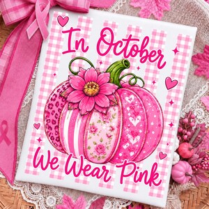 Op de afbeelding: Een rechthoekig bord met een roze en witte gingham achtergrond en een grote roze pompoen met bloemen-, luipaard- en hartjespatronen. Bovenop zit een roze bloem. De tekst In October We Wear Pink staat in roze cursief geschreven.