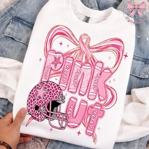 Könnte beinhalten: Weißes Sweatshirt mit einem rosa und goldenen Design. Das Design umfasst die Worte "PINK OUT" in rosa Football-Schrift, einen rosa Footballhelm mit Leopardenmuster und ein rosa Band. Das Sweatshirt befindet sich auf einem Jeans-Hintergrund.
