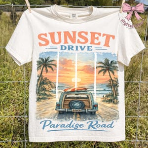 May include: Off-white t-shirt with a graphic of a vintage car on a beach road at sunset. The text "Sunset Drive" and "Paradise Road" are printed above and below the image. Palm trees and a surfboard are also visible.