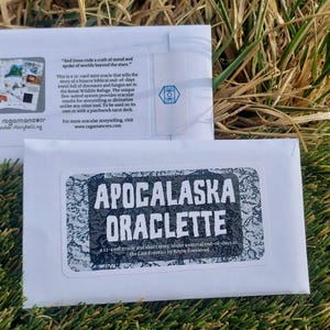 Puede incluir: Dos sobres blancos con texto y gráficos se muestran sobre hierba. El sobre inferior presenta una pegatina con las palabras "APOCALASKA ORACLETTE". El sobre superior tiene texto y un logotipo que dice "the ragamancer". La imagen está tomada al aire libre.