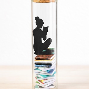 May include: A clear glass bottle with a cork stopper, containing a stack of miniature books. A black silhouette of a person reading sits atop the books. The text "Tiny Library in a Bottle" is at the bottom.