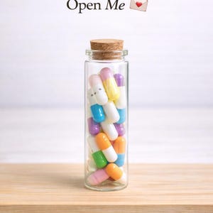May include: A clear glass bottle filled with colorful capsules, topped with a cork. The capsules are various colors, including pink, orange, blue, and yellow. The words "Open Me" are above the bottle, with a small heart envelope icon.