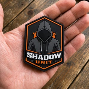 Könnte beinhalten: Ein sechseckiger Aufnäher mit einer dunkelgrauen Kapuzenfigur vor einem orangefarbenen kreisförmigen Hintergrund. Der Text SHADOW UNIT ist in weißen und orangefarbenen Blockbuchstaben unten aufgedruckt, flankiert von zwei kleinen Sternen.