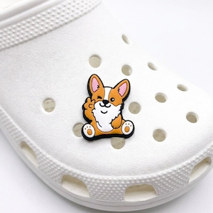 May include: A white foam clog shoe featuring a decorative rubber charm shaped like a cartoon corgi dog. The corgi is orange and white, winking with one eye and holding up a paw in a peace sign gesture, attached to the top of the footwear.