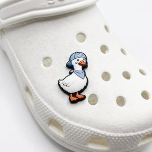 Op de afbeelding: Een witte rubberen schoenbedel in de vorm van een cartoon-eend die een lichtblauwe honkbalpet en een bijpassende lichtblauwe halsdoek draagt. De eend heeft een wit lichaam, een oranje snavel en oranje voeten, bevestigd op een witte schoen.