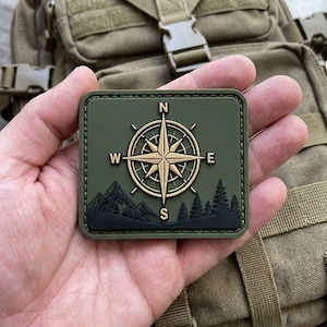 May include: A square olive green tactical patch featuring a raised gold compass rose in the centre with cardinal directions N, E, W, and S. Below the compass is a black silhouette of a mountain range and a forest of pine trees.