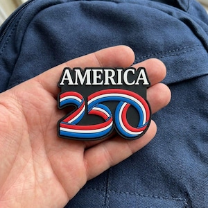 May include: A black rubber patch featuring the white text AMERICA above the number 250. The number 250 is stylised with a ribbon design in red, white, and blue stripes that curve to form the digits, set against a solid black background.