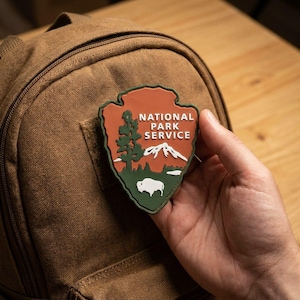 Può includere: Una toppa a forma di scudo del National Park Service con una parte superiore color arancio terracotta con testo bianco, una sezione inferiore verde scuro con un albero bianco, una catena montuosa bianca e la sagoma di un bisonte bianco su uno zaino in tela marrone.