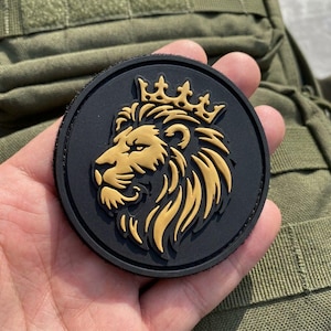Pode incluir: Um emblema circular preto com uma cabeça de leão dourada em relevo, de perfil virada para a esquerda, encimada por uma coroa estilizada. O design é moldado com linhas nítidas e um acabamento mate, criando um contraste entre o dourado e o preto.