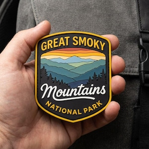 May include: A shield-shaped rubber patch featuring a graphic of the Great Smoky Mountains. The design includes a sunset sky, layered blue mountain ranges, and black pine trees. The text reads GREAT SMOKY MOUNTAINS NATIONAL PARK in yellow.