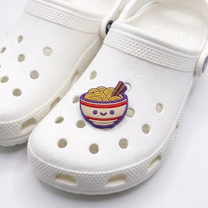 Può includere: Uno zoccolo in schiuma bianco con un ciondolo decorativo a forma di ciotola di ramen. Il ciondolo presenta un volto sorridente su una ciotola color crema con strisce rosse e viola, contenente noodles gialli e bacchette marroni.