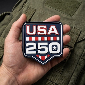 Op de afbeelding: Een rechthoekige rubberen patch met afgeronde hoeken en een puntige onderkant. De patch heeft een donkerblauwe achtergrond met witte tekst USA 250, geaccentueerd door rode en witte horizontale strepen en een kleine witte ster in de nul.