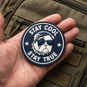 Pode incluir: Um emblema circular azul-marinho com um gráfico branco de um bulldog usando óculos de sol pretos e uma coleira com espinhos. O texto "STAY COOL" está arqueado acima e "STAY TRUE" abaixo, com duas estrelas brancas nas laterais.