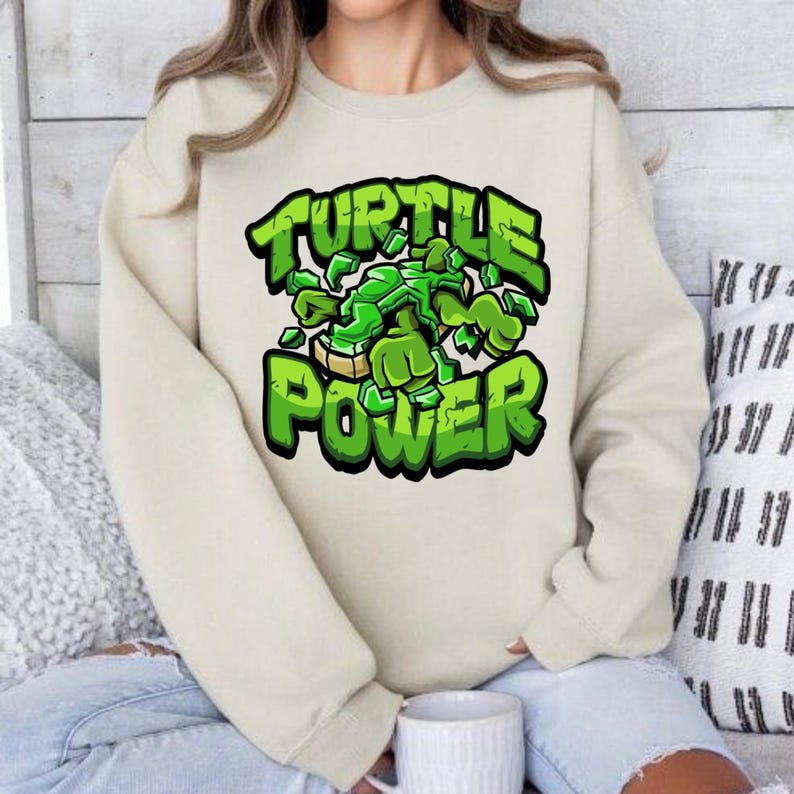 Pode incluir: Um moletom creme com um gr&aacute;fico verde de uma tartaruga e as palavras "TURTLE POWER" em negrito, verde. O design tem um estilo de desenho animado, com um efeito 3D. O moletom tem mangas compridas e gola redonda.