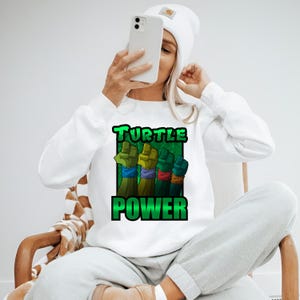 Pode incluir: Camisola branca com um gr&aacute;fico com quatro punhos em verde, amarelo, verde e azul, cada um com uma pulseira de cor diferente. As palavras "TURTLE POWER" est&atilde;o impressas em verde. A camisola &eacute; usada por uma pessoa.