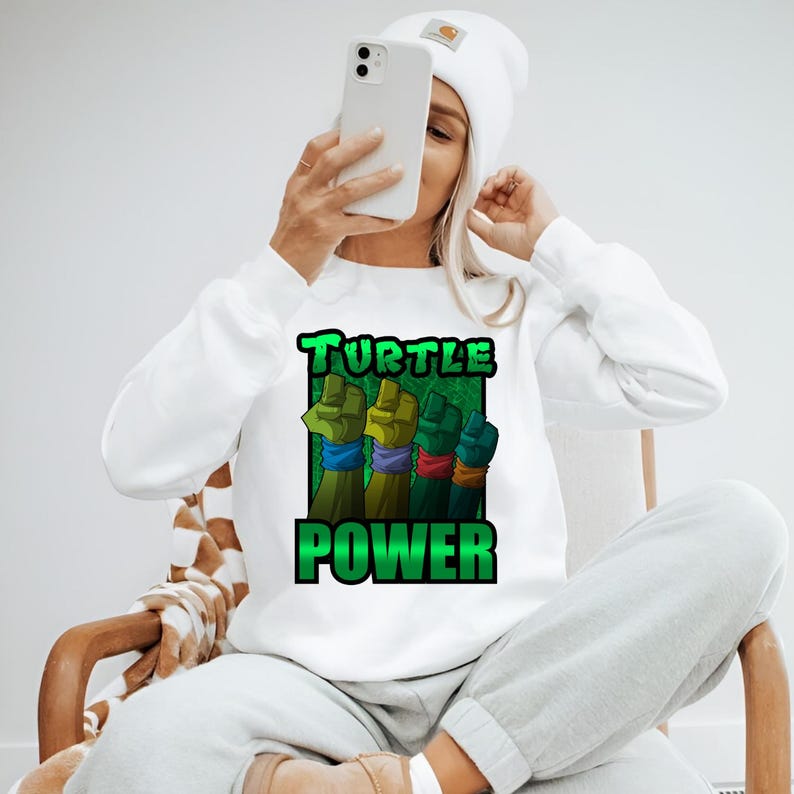 Pode incluir: Camisola branca com um gr&aacute;fico com quatro punhos em verde, amarelo, verde e azul, cada um com uma pulseira de cor diferente. As palavras "TURTLE POWER" est&atilde;o impressas em verde. A camisola &eacute; usada por uma pessoa.