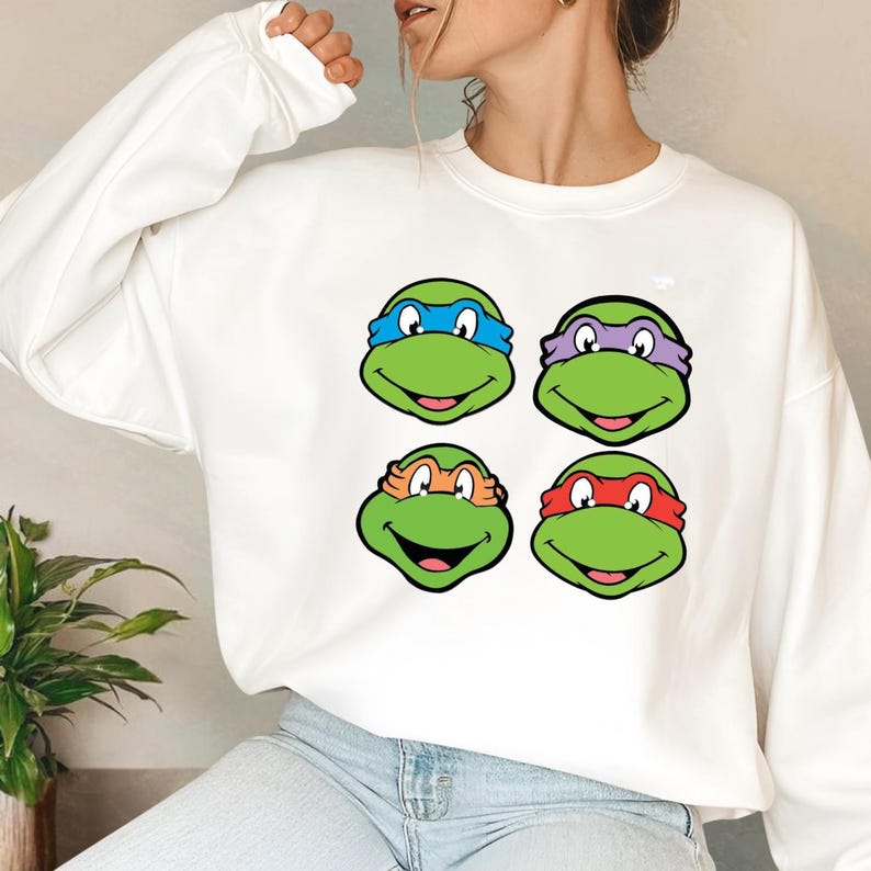 Pode incluir: Camisola branca com um desenho de quatro rostos de tartarugas de desenho animado, cada um com uma bandana de cor diferente: azul, roxo, laranja e vermelho. As tartarugas t&ecirc;m rostos verdes e express&otilde;es sorridentes. Um estilo casual e confort&aacute;vel.