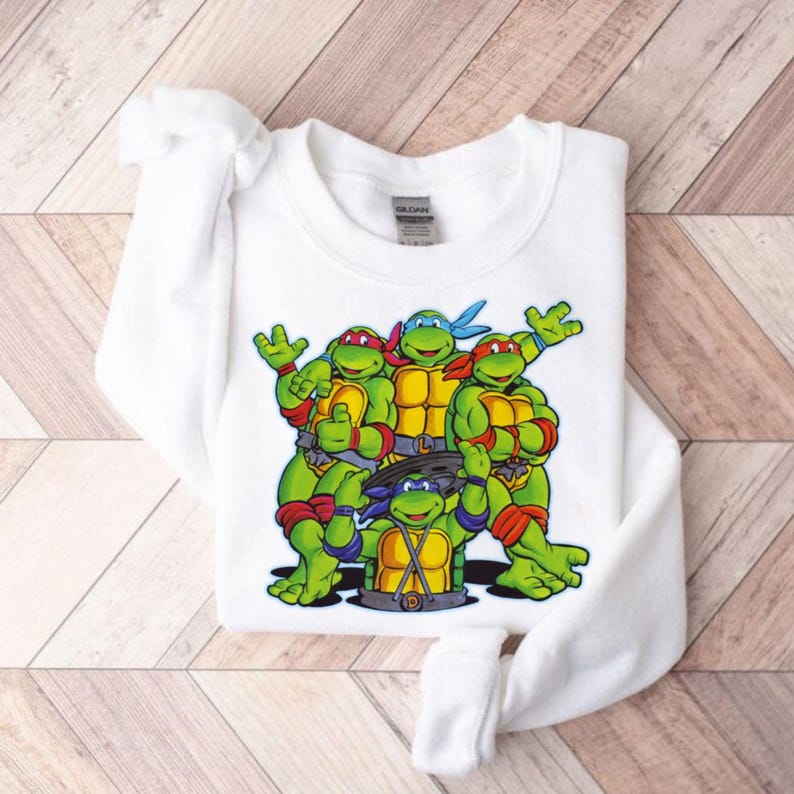 Pode incluir: Camisola branca com um gr&aacute;fico das Tartarugas Ninja. As tartarugas s&atilde;o verdes com m&aacute;scaras e carapa&ccedil;as coloridas, posando em grupo. A camisola est&aacute; dobrada sobre uma superf&iacute;cie clara com gr&atilde;o de madeira.