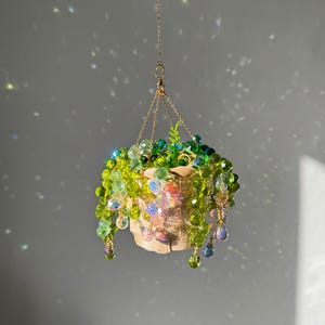 May include: A hanging basket with a woven design, filled with vibrant green and yellow beaded foliage. The beads cascade down, creating a decorative, plant-like appearance. The basket is suspended by a delicate gold chain, adding a touch of elegance to this unique home decor item.