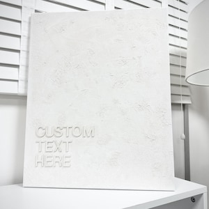 May include: A textured white canvas with the words "CUSTOM TEXT HERE" embossed on the front. The canvas is rectangular and stands on a white shelf. The background includes white blinds and a lamp.