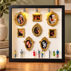 May include: A black shadow box frame containing seven ornate gold-coloured picture frames arranged on a white background. Three small lights are mounted at the top. Miniature figures of people are standing along the bottom edge of the display.