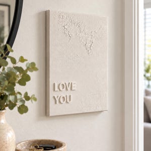 May include: A rectangular wall art piece in a neutral beige tone featuring a textured, cracked surface finish on the upper portion and the raised, three-dimensional lettering "LOVE YOU" in a clean, sans-serif font on the lower left side.