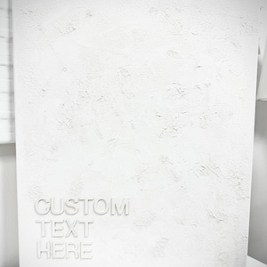 May include: A rectangular white canvas featuring a heavily textured, impasto-style surface. The lower left corner displays the embossed, raised white lettering that reads CUSTOM TEXT HERE in a clean, sans-serif font against the rough background.