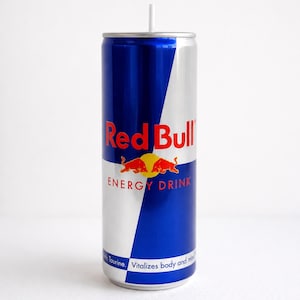 May include: A tall, cylindrical aluminium energy drink can featuring a blue and silver colour scheme. The Red Bull logo is printed in red and yellow, with the text Red Bull Energy Drink and the phrase Vitalizes body and mind visible.
