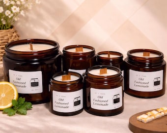 May include: A collection of brown glass jar candles with wooden wicks. Each candle has a white label that reads "Old Fashioned Lemonade" with a small illustration. A sliced lemon and greenery are in the foreground.