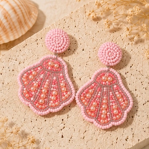 May include: A pair of dangle earrings featuring a circular stud top and a large shell-shaped drop, both covered in pink seed beads. The shell design incorporates rows of small, round, cream-colored faux pearls and silver metallic thread accents.