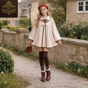 May include: A cream-colored cape with brown trim and a matching collar. The cape features two decorative pom-poms and is paired with brown boots and a red beret. The Regency Store logo is visible in the upper left corner.