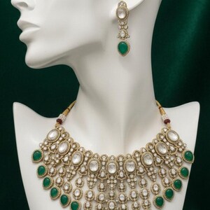 Op de afbeelding: Een goudkleurige ketting en bijpassende oorbellen set met heldere en smaragdgroene edelstenen. De ketting heeft een slabbetje ontwerp met meerdere rijen stenen, weergegeven op een witte mannequin hoofd tegen een donkergroene achtergrond.