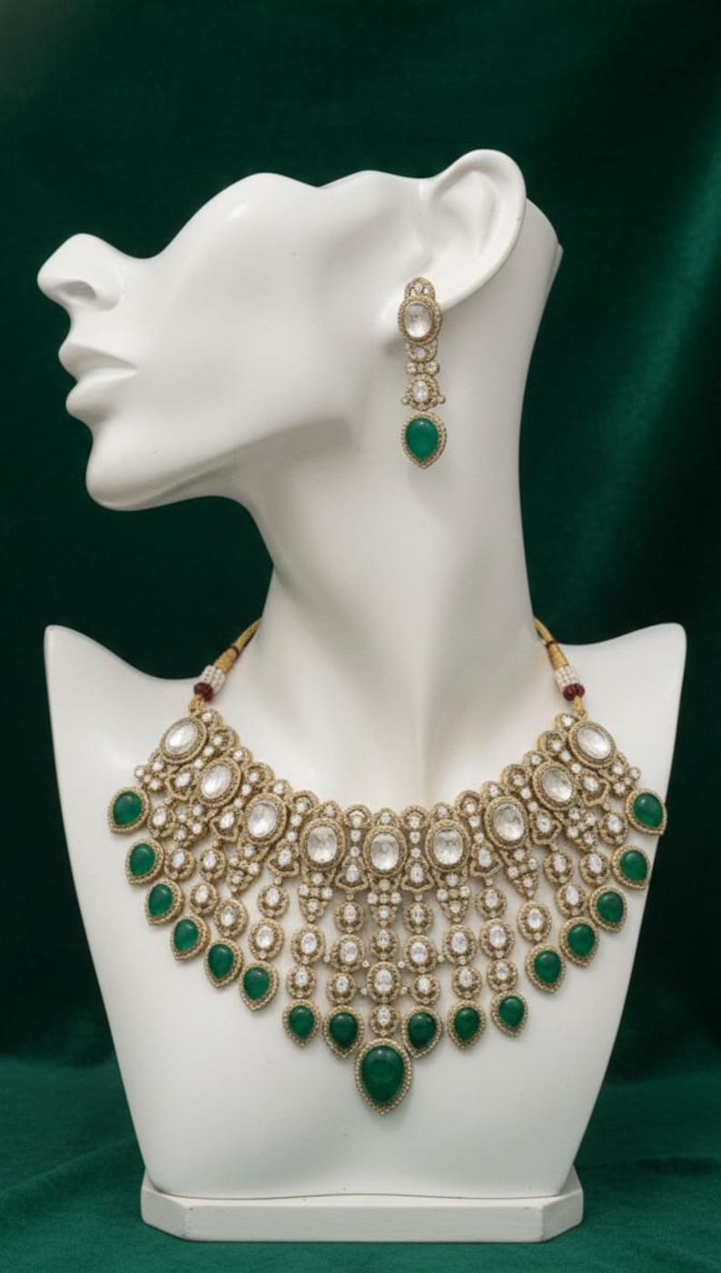 Op de afbeelding: Een goudkleurige ketting en bijpassende oorbellen set met heldere en smaragdgroene edelstenen. De ketting heeft een slabbetje ontwerp met meerdere rijen stenen, weergegeven op een witte mannequin hoofd tegen een donkergroene achtergrond.